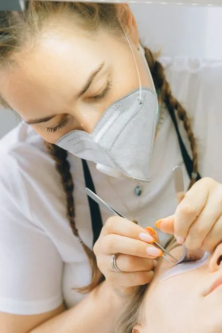 Technicienne masquée travaillant à la pince sur les cils d'une cliente allongée.